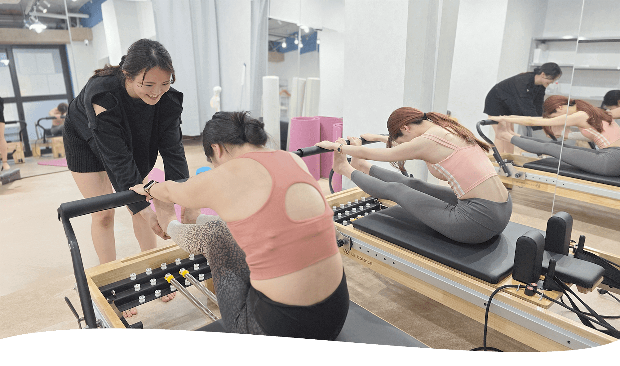 女性二人がマシンの上で前屈になるストレッチを一人の女性のインストラクターの方に指導されている画像